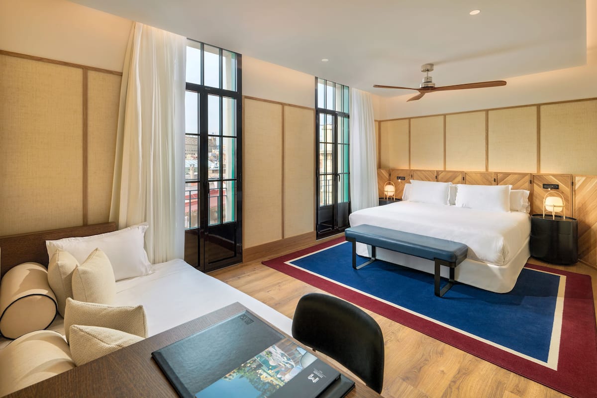 Ein Hotelzimmer mit einem großen Bett, einem Schreibtisch mit Stuhl, einer Bank und Blick auf die Stadt durch große Fenster.
