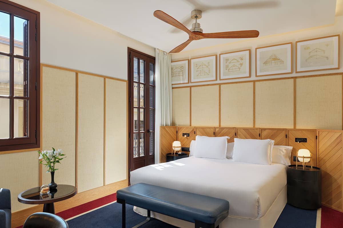 Ein Hotelzimmer mit einem großen Bett, einem Holz-Kopfteil, einem Deckenventilator, einem kleinen Tisch mit Blumenvase und gerahmten architektonischen Zeichnungen an der Wand.