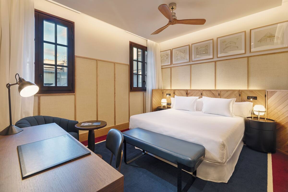 A hotel room with a bed, bedside tables, a desk, a chair, a bench, a ceiling fan, and framed artwork on the wall.