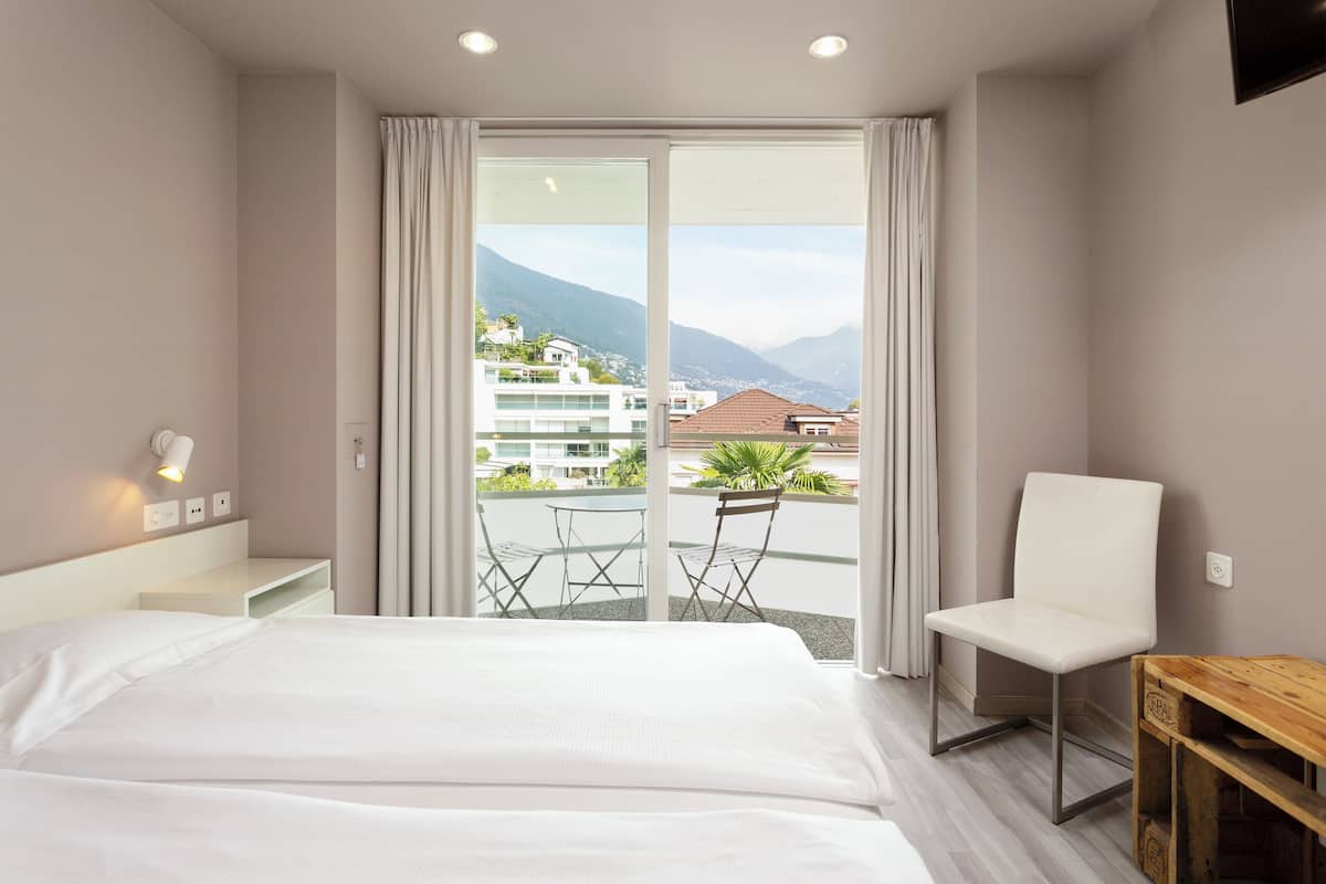 A hotel room with a large bed, a chair, a small table, and a balcony with a view of buildings and mountains.