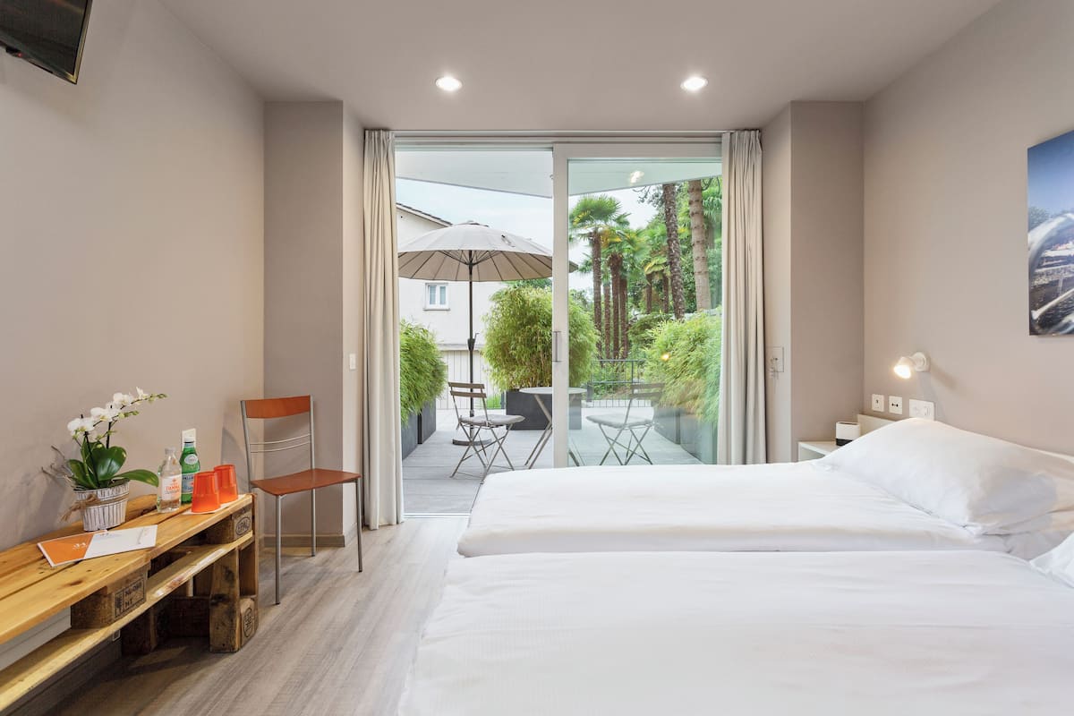 A hotel room with a bed, a desk with a chair, a small table with a plant, and a view of an outdoor patio with a table and chairs.
