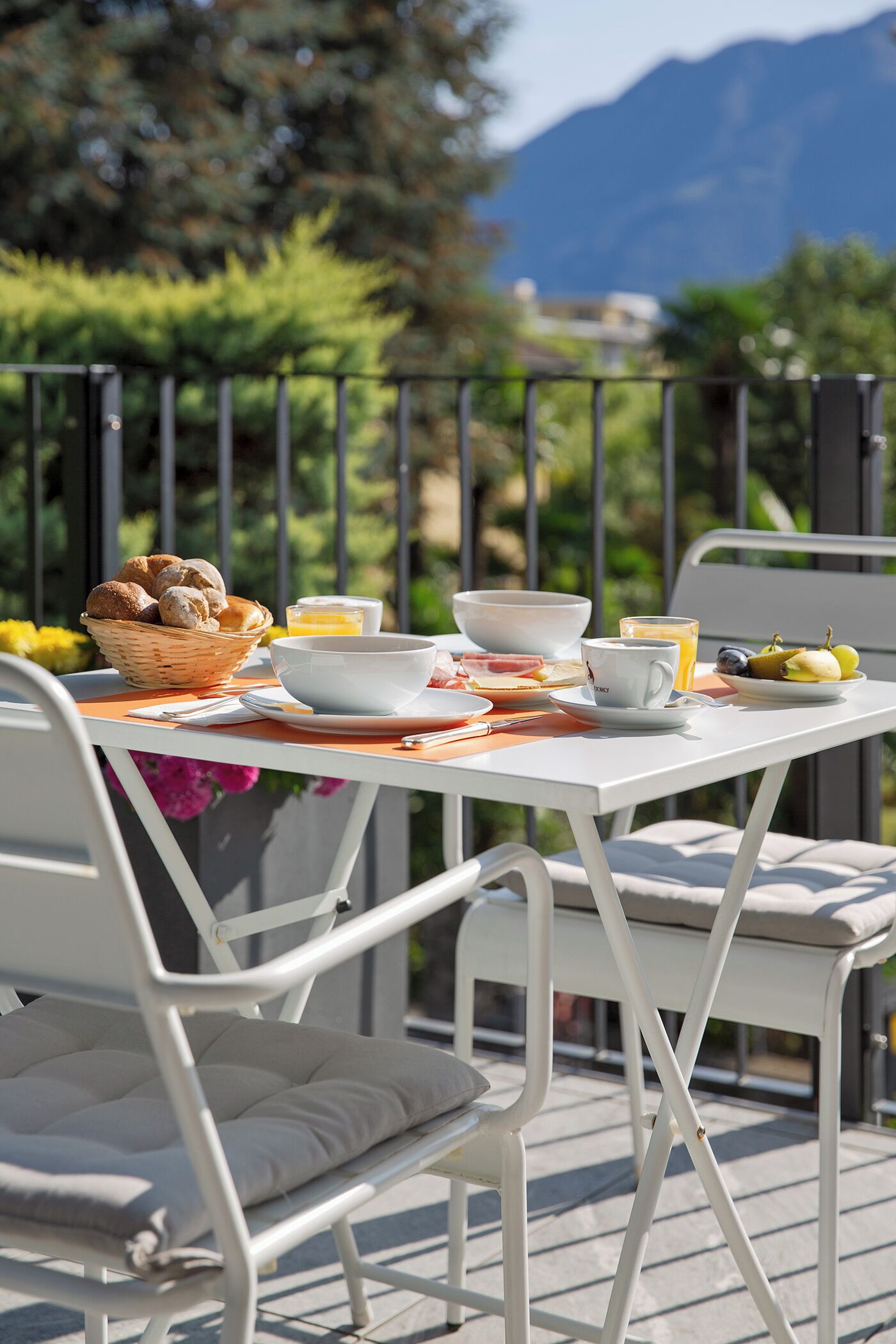 A breakfast table set outdoors with a basket of bread, a bowl, a cup, and a plate of fruit.