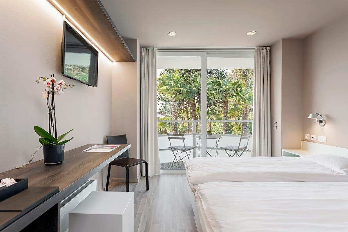 A modern hotel room with a bed, desk, chair, and a balcony with a view of palm trees.