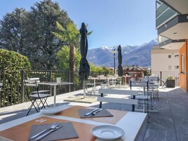 Un espacio de comedor al aire libre con mesas y sillas, vistas a montañas y un edificio con balcones.