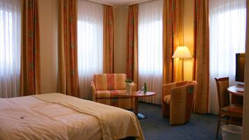 Habitación de hotel con cama, sofá, sillón, una mesita con lámpara y televisor.