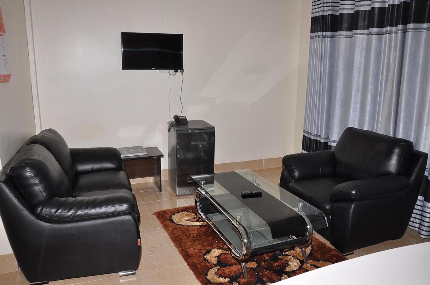 A living room with two black leather sofas, a glass coffee table, a TV mounted on the wall, and a small side table with a lamp.
