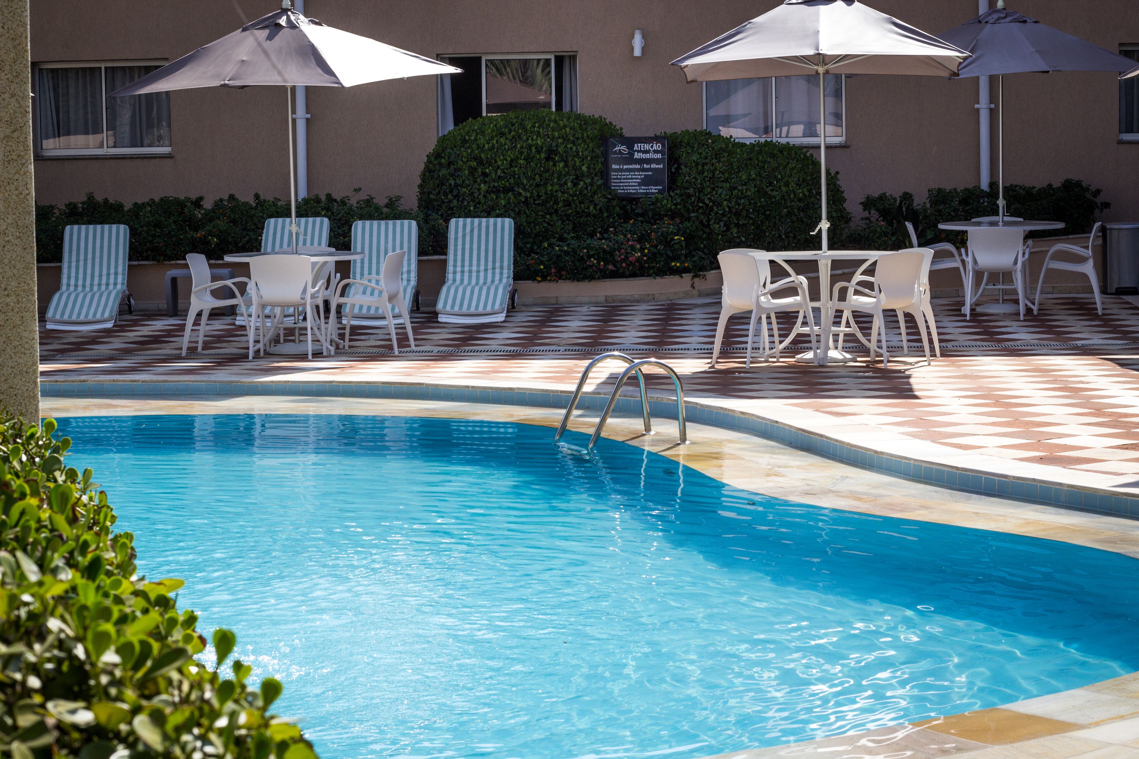 Zona de piscina con sillas de descanso y sombrillas, piscina con escalera y un cartel que indica atención a los huéspedes.