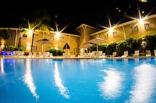 Una piscina nocturna con sillones de descanso y sombrillas, rodeada por un edificio con múltiples ventanas y balcones.