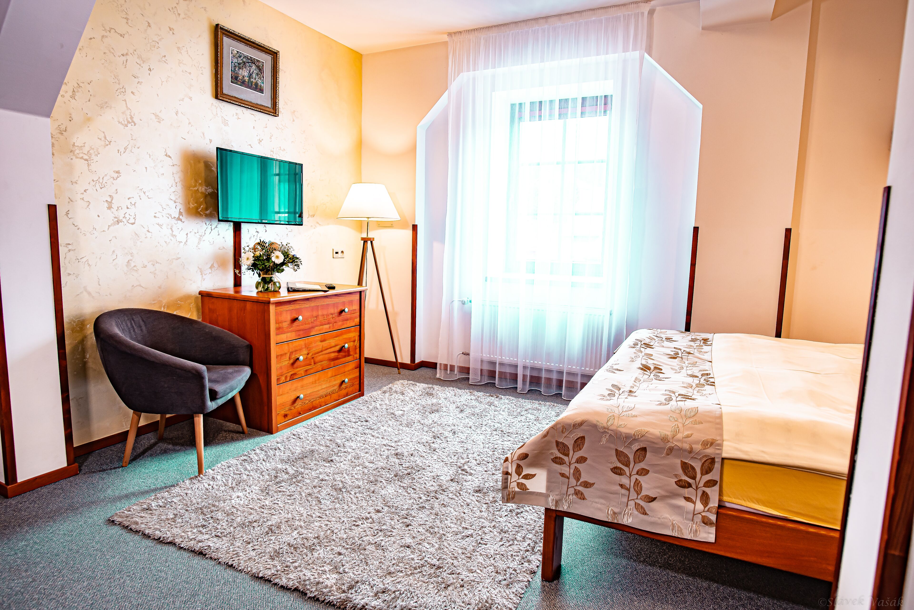 Ein Schlafzimmer mit Bett, Schreibtisch, Stuhl, Lampe und einem Fenster mit Vorhängen.