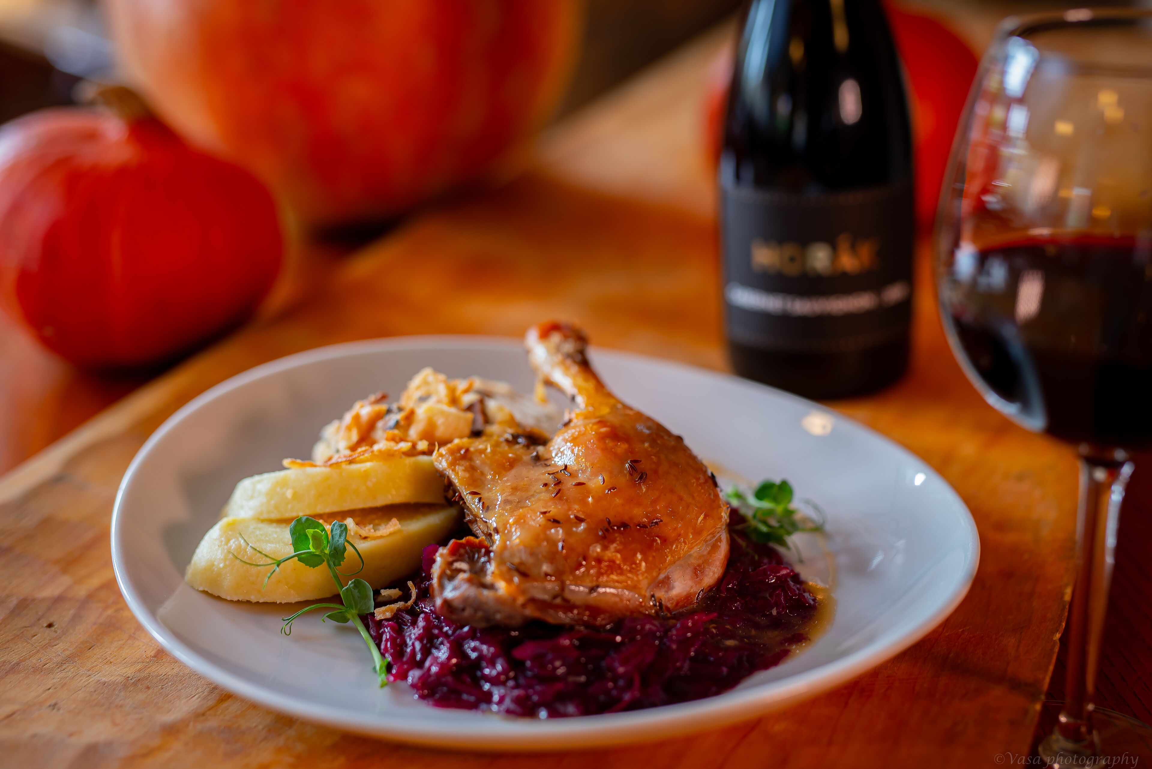 A plate with roasted chicken, potatoes, and red cabbage, accompanied by a bottle of wine and a glass of red wine on a wooden table.