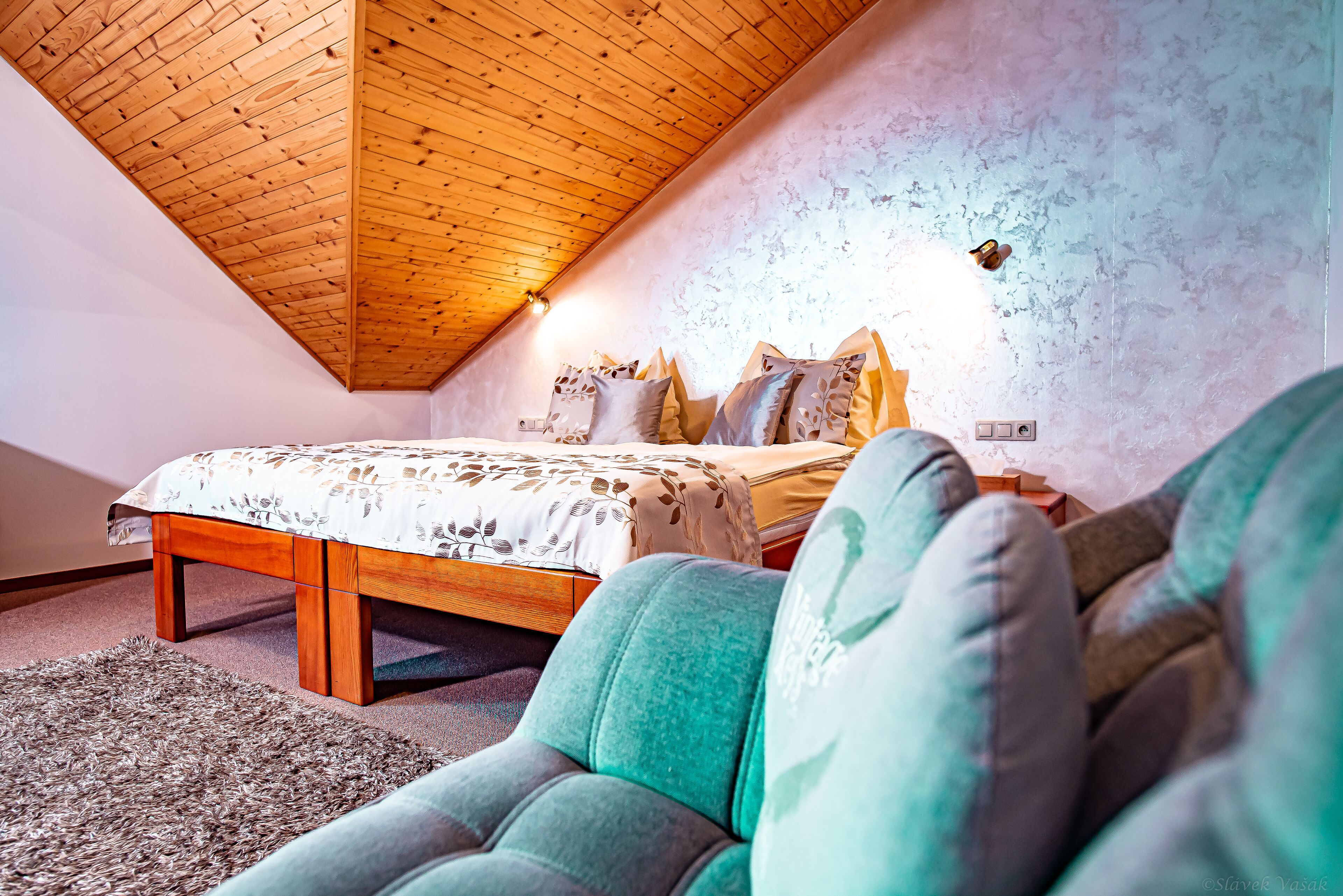 A bedroom with a wooden ceiling, a bed with patterned bedding, and a green armchair.