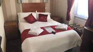A hotel room with a neatly made bed, a wooden nightstand, a red throw blanket, and a folded towel on the bed.