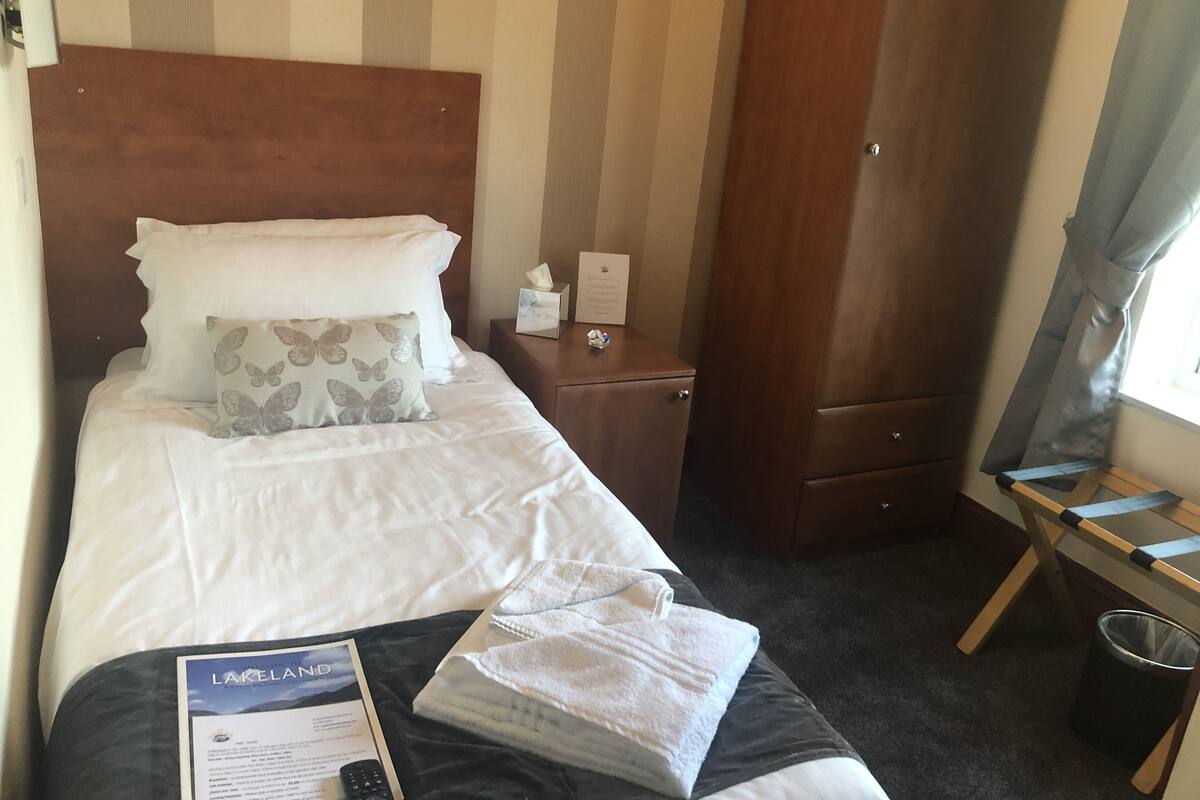 Quarto de hotel com cama, mesa de cabeceira, guarda-roupa de madeira, janela com cortinas e pequena mesa com toalha e controle remoto.