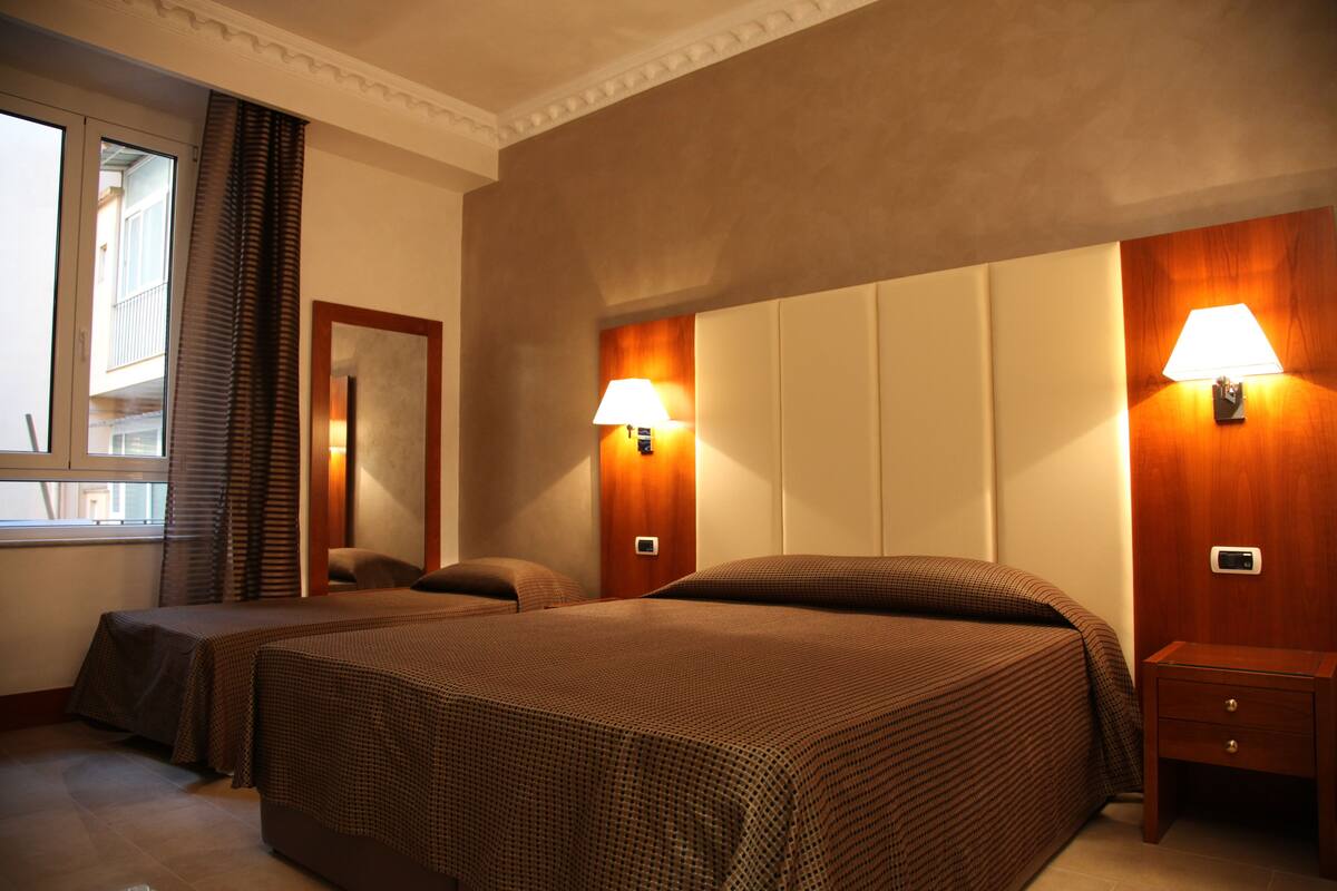 Ein Hotelzimmer mit zwei Betten, einem Nachttisch, einer Lampe und einem Fenster mit Vorhängen.