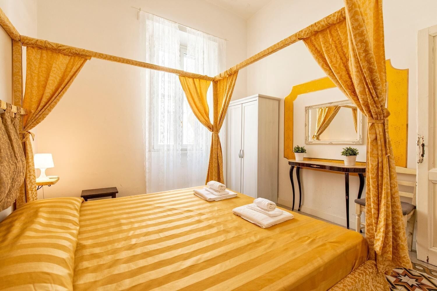 A room with a four-poster bed, a bedside table with a lamp, a mirror, and a small potted plant.