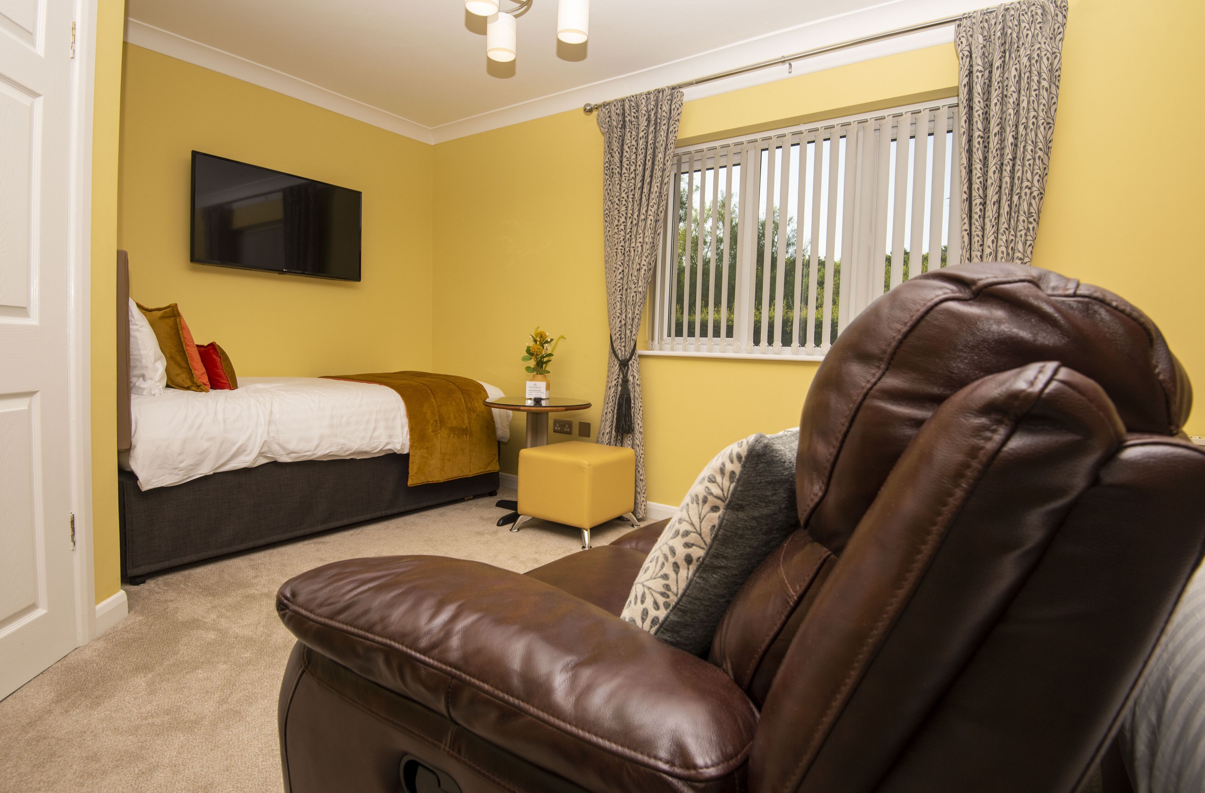 A hotel room with a bed, a sofa, a small table with a vase, and a window with curtains.