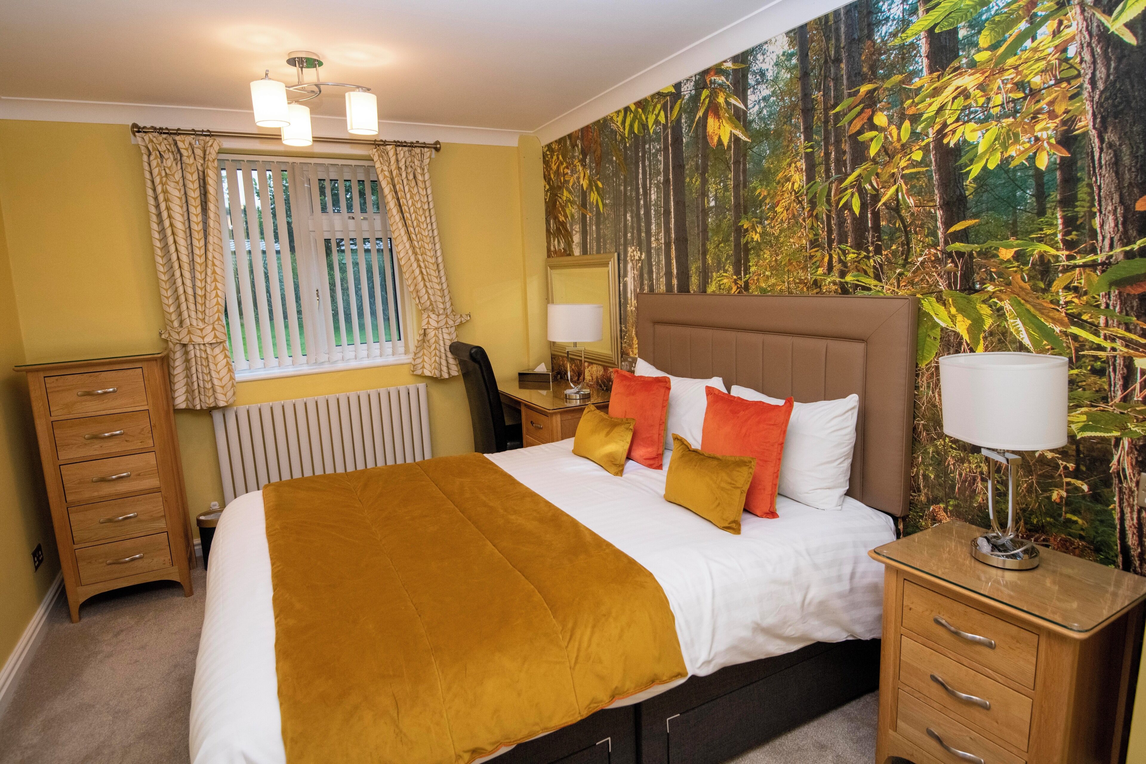 Hotelkamer met een bed, nachtkastjes, een kaptafel, een raam met gordijnen, een radiator en een behang met bosmotief.