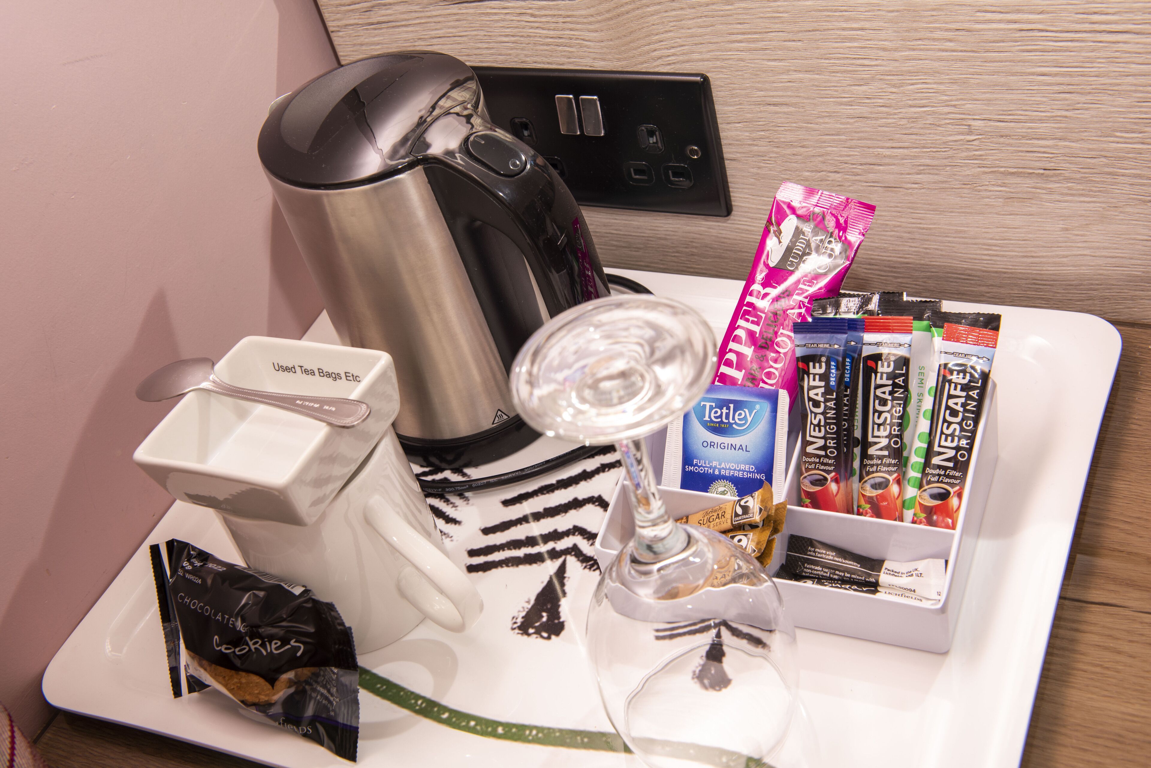 Una bandeja con una cafetera, un vaso de agua, un dispensador de bolsitas de té y una caja de sobres de café instantáneo Nescafé.