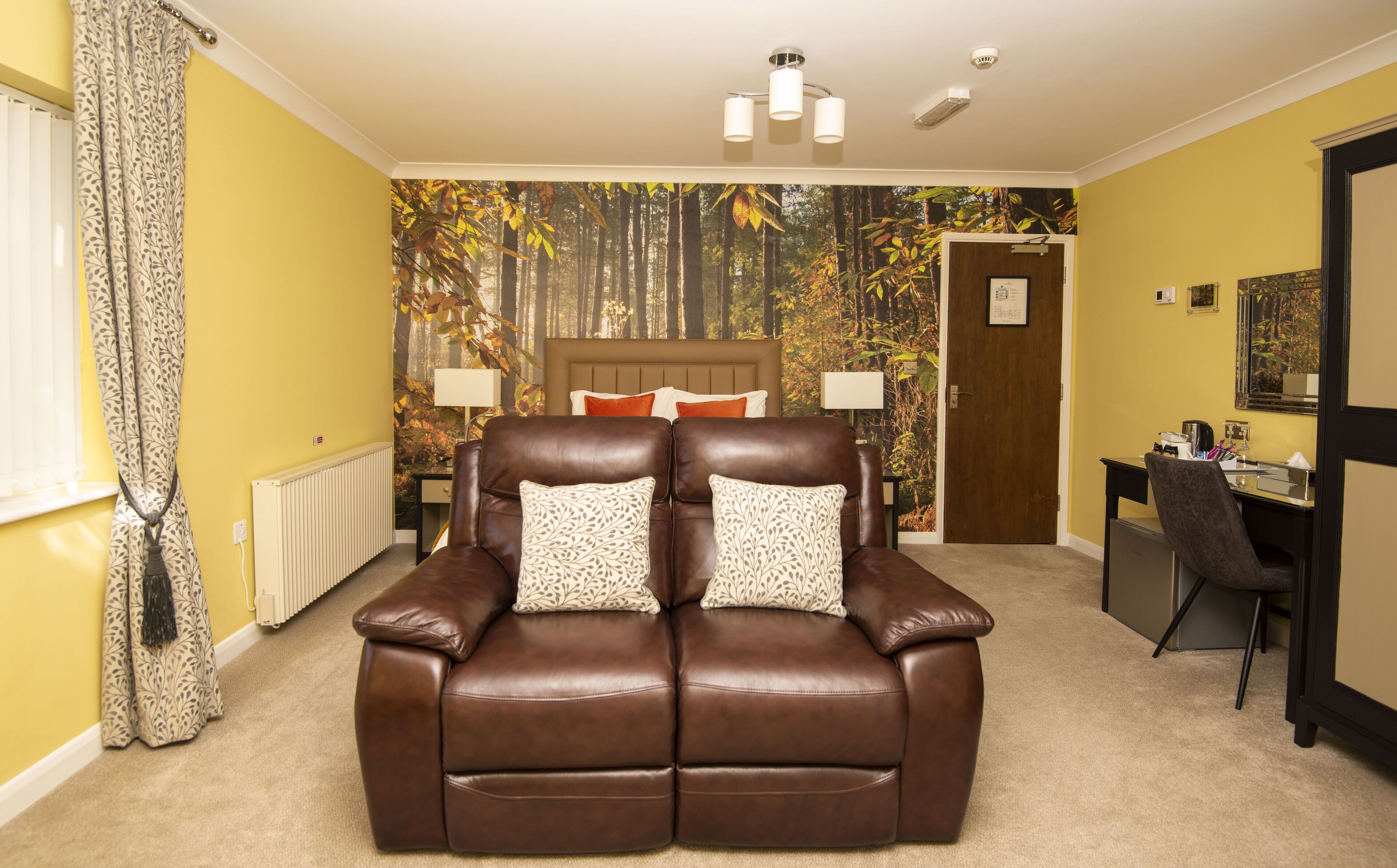 A room with a brown leather sofa, a desk, and a wall with a forest mural.