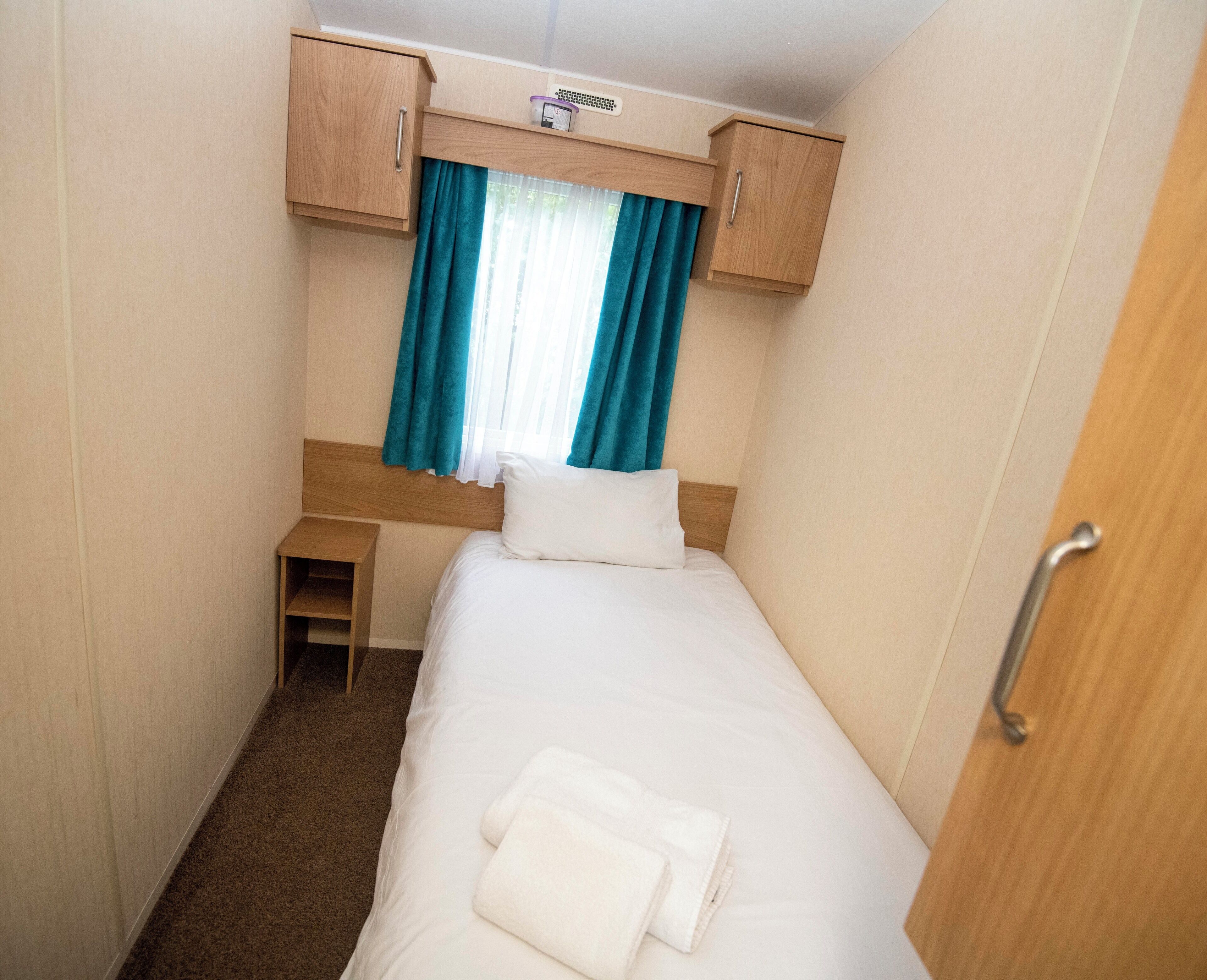 A compact bedroom with a single bed, a small bedside table, a wooden wardrobe, and a window with curtains.