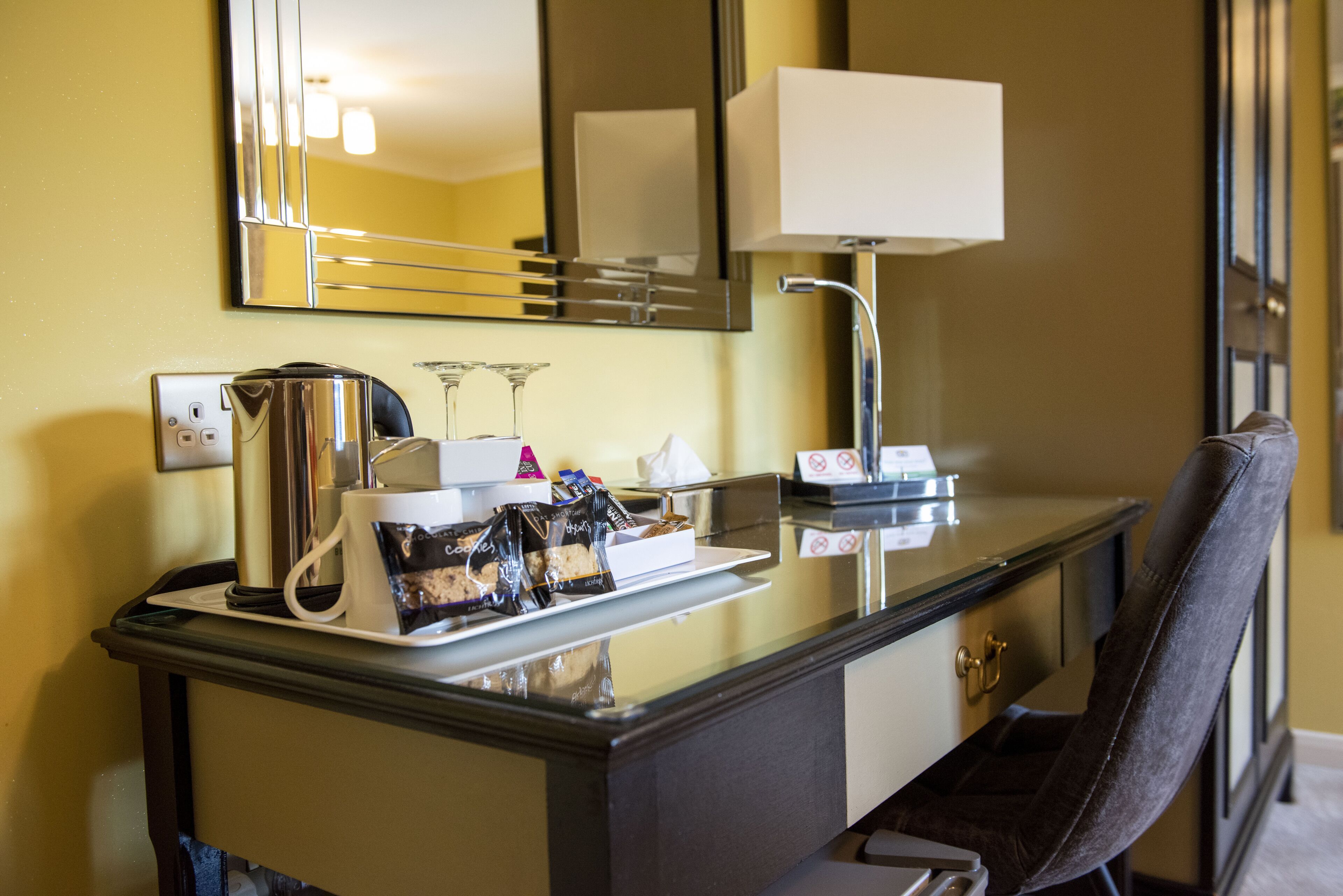 A hotel desk with a coffee maker, a tray of snacks, a mirror, and a lamp.