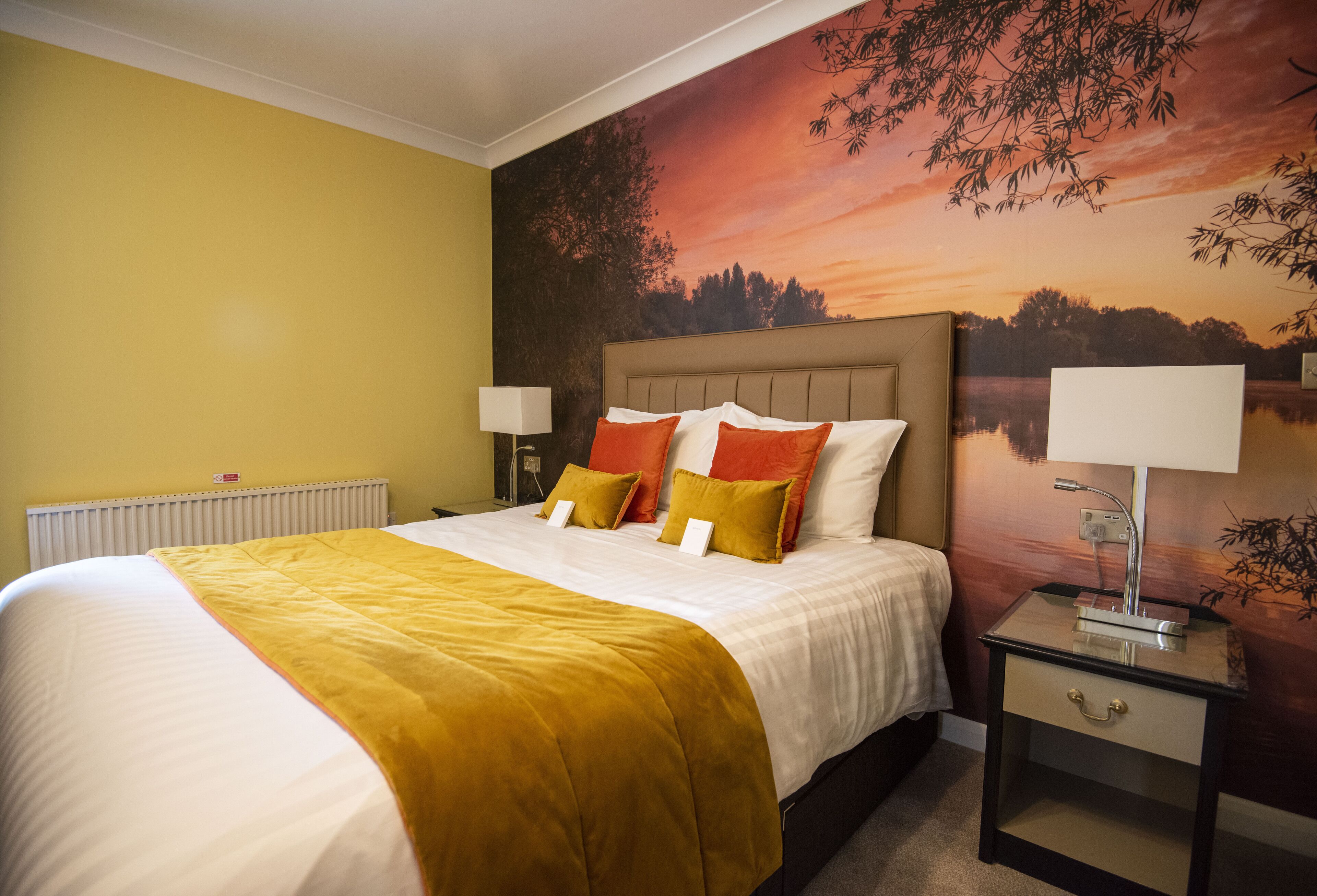 Habitación de hotel con una cama grande, una mesita de noche con lámpara y un mural en la pared que representa un atardecer sobre un lago.