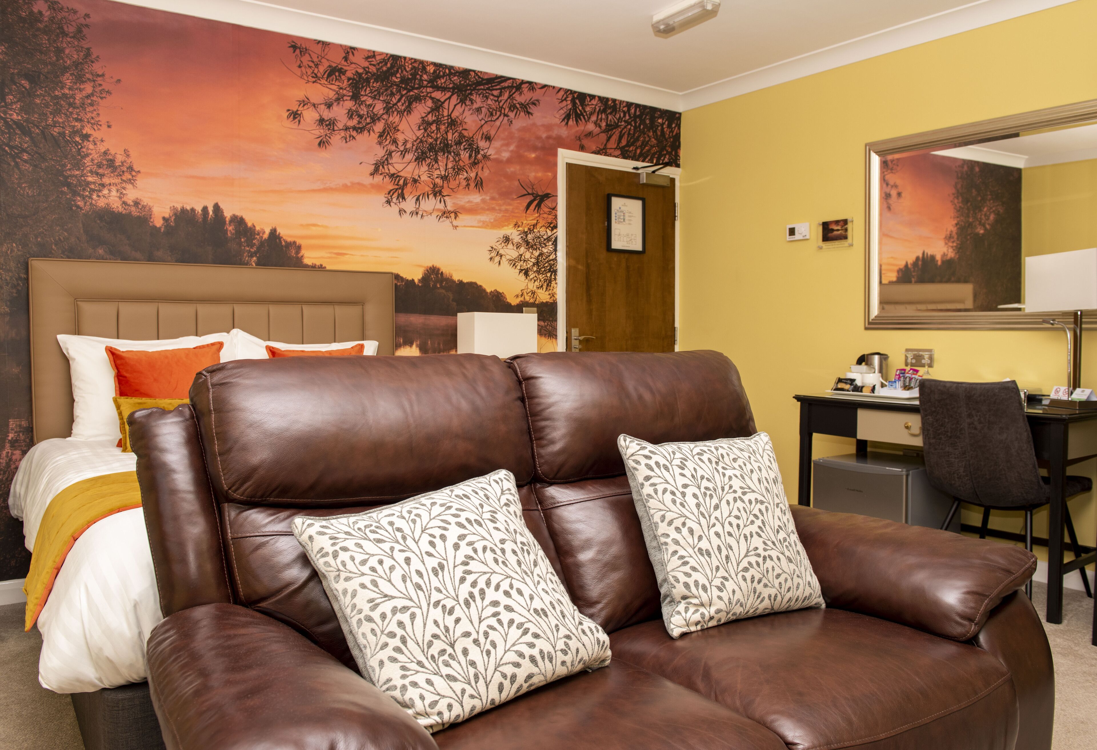 A room with a brown leather sofa, a bed with orange and white bedding, a desk with a chair, and a large mirror on the wall.