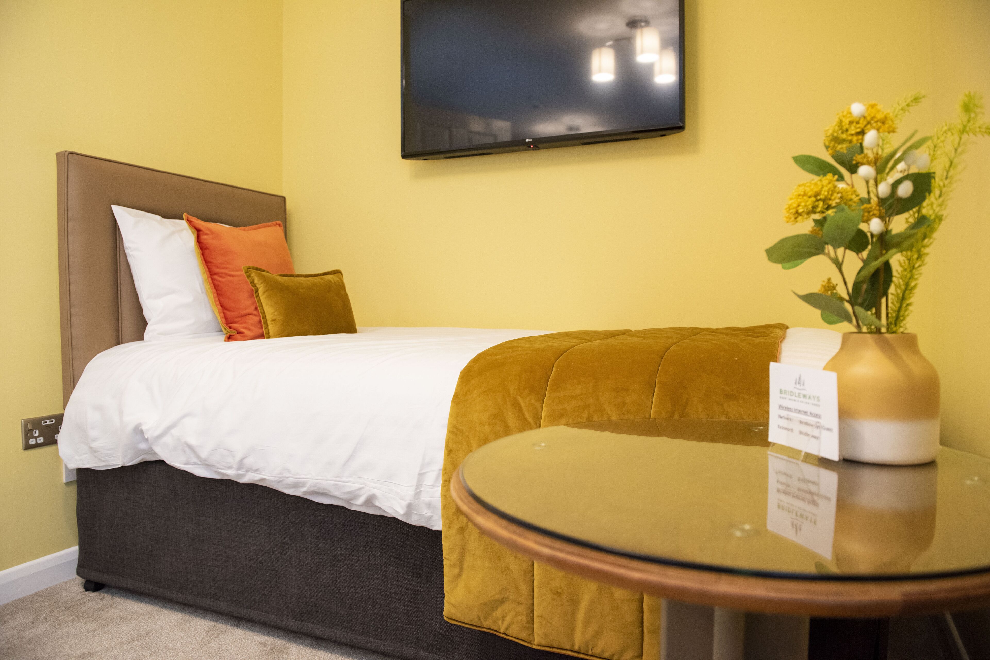 A hotel room with a bed, a round glass table, a TV mounted on the wall, and a vase with flowers on a side table.
