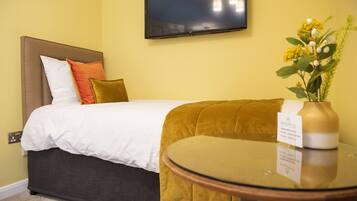 A hotel room with a bed, a round glass table, a TV mounted on the wall, and a vase with flowers on a side table.
