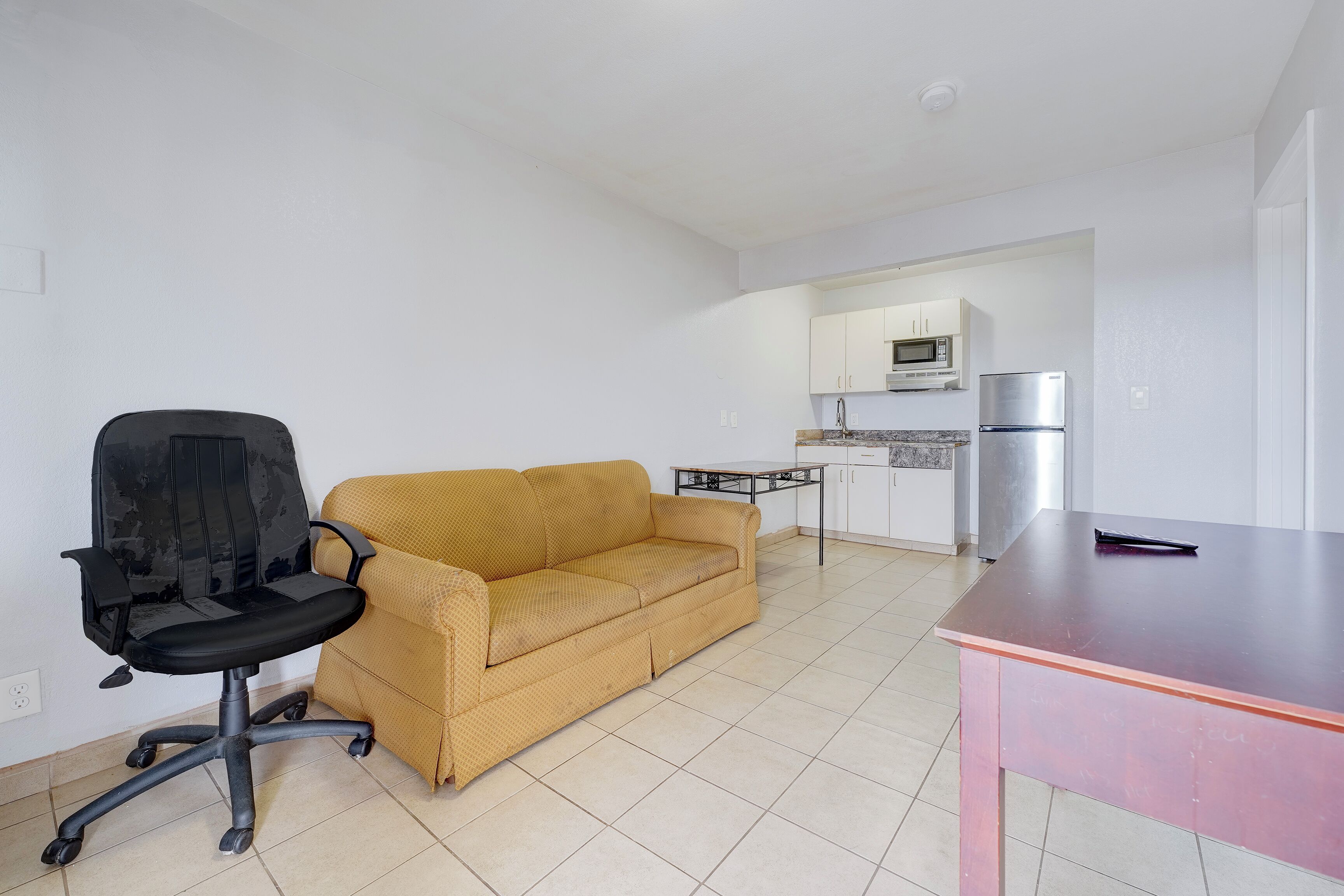 Una sala de estar con un sofá amarillo, una silla de oficina negra, una mesita y una cocina al fondo.