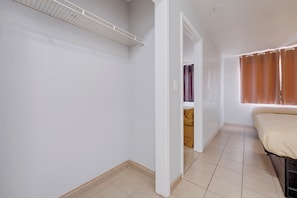 A bedroom with a bed, a window with curtains, a door leading to another room, and a tiled floor.