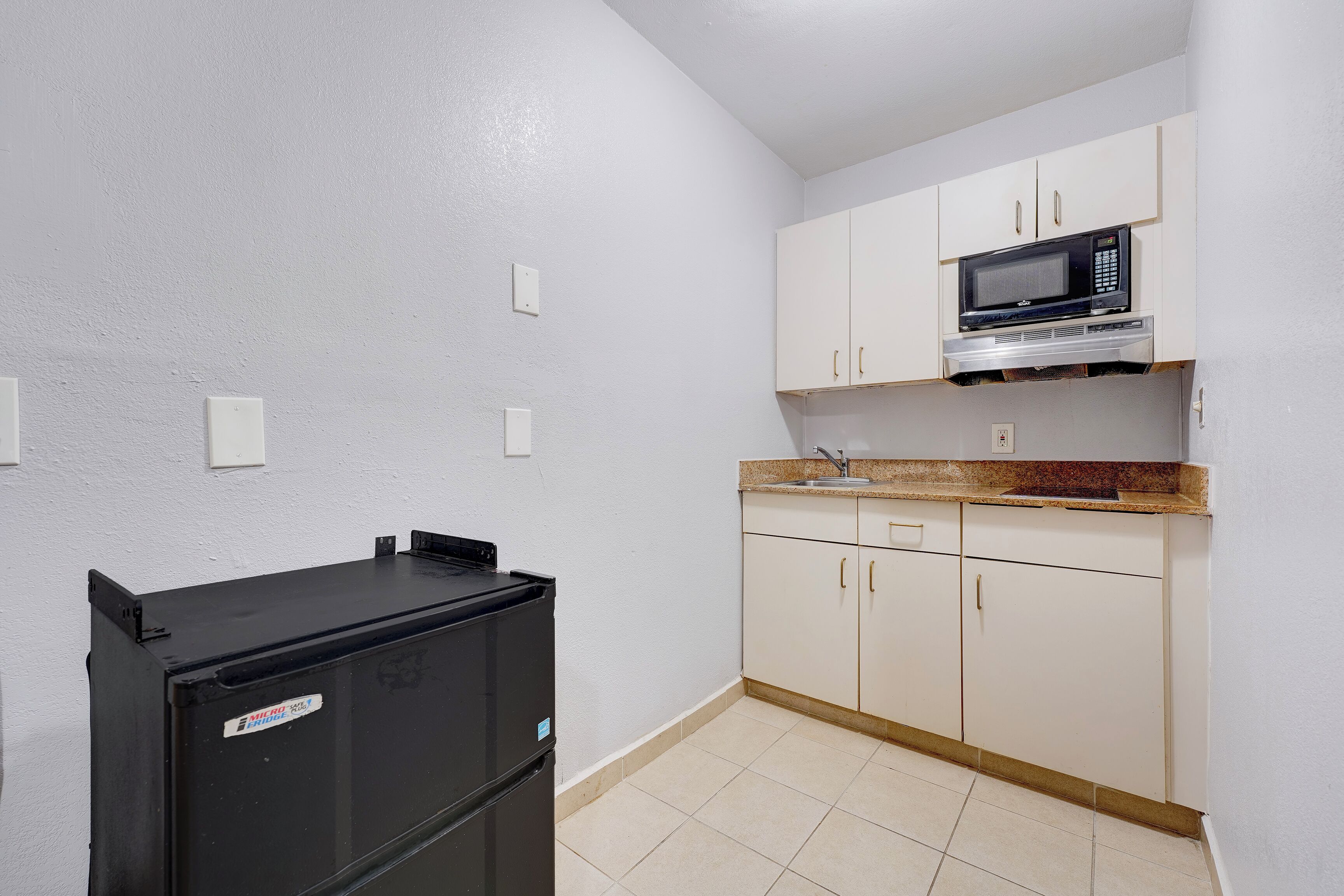 Cocina compacta con armarios blancos, un refrigerador negro y un microondas.