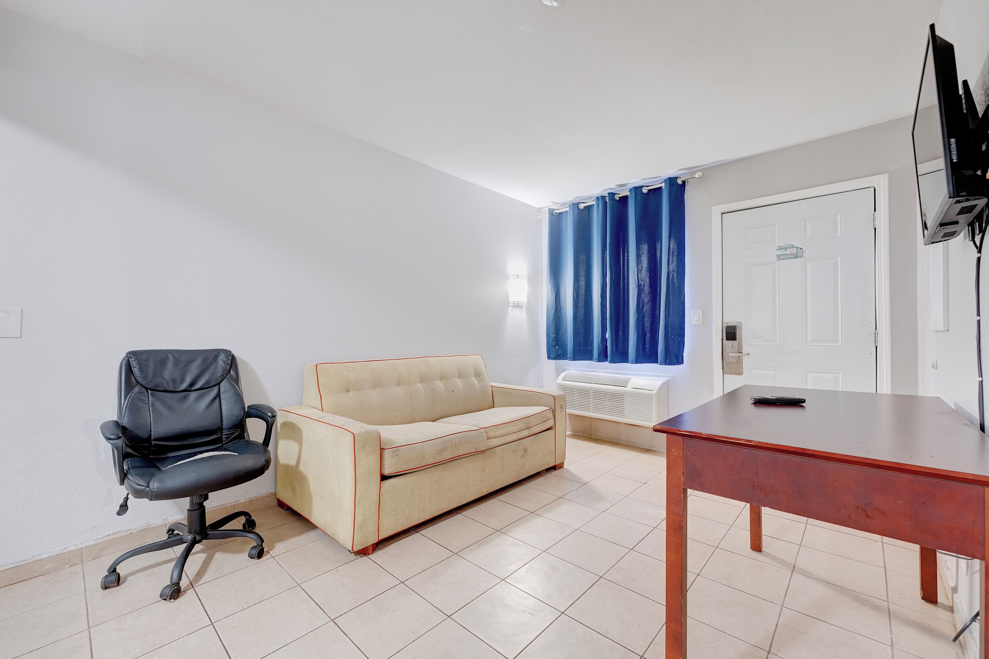 A room with a black office chair, a beige sofa, a wooden table, and a TV on a stand.