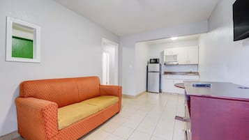 Una habitación con sofá, una zona de cocina con armarios y electrodomésticos, y un televisor montado en la pared.