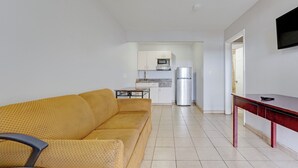 A compact living space with a sofa, a small table, a kitchen area with a microwave and refrigerator, and a TV mounted on the wall.