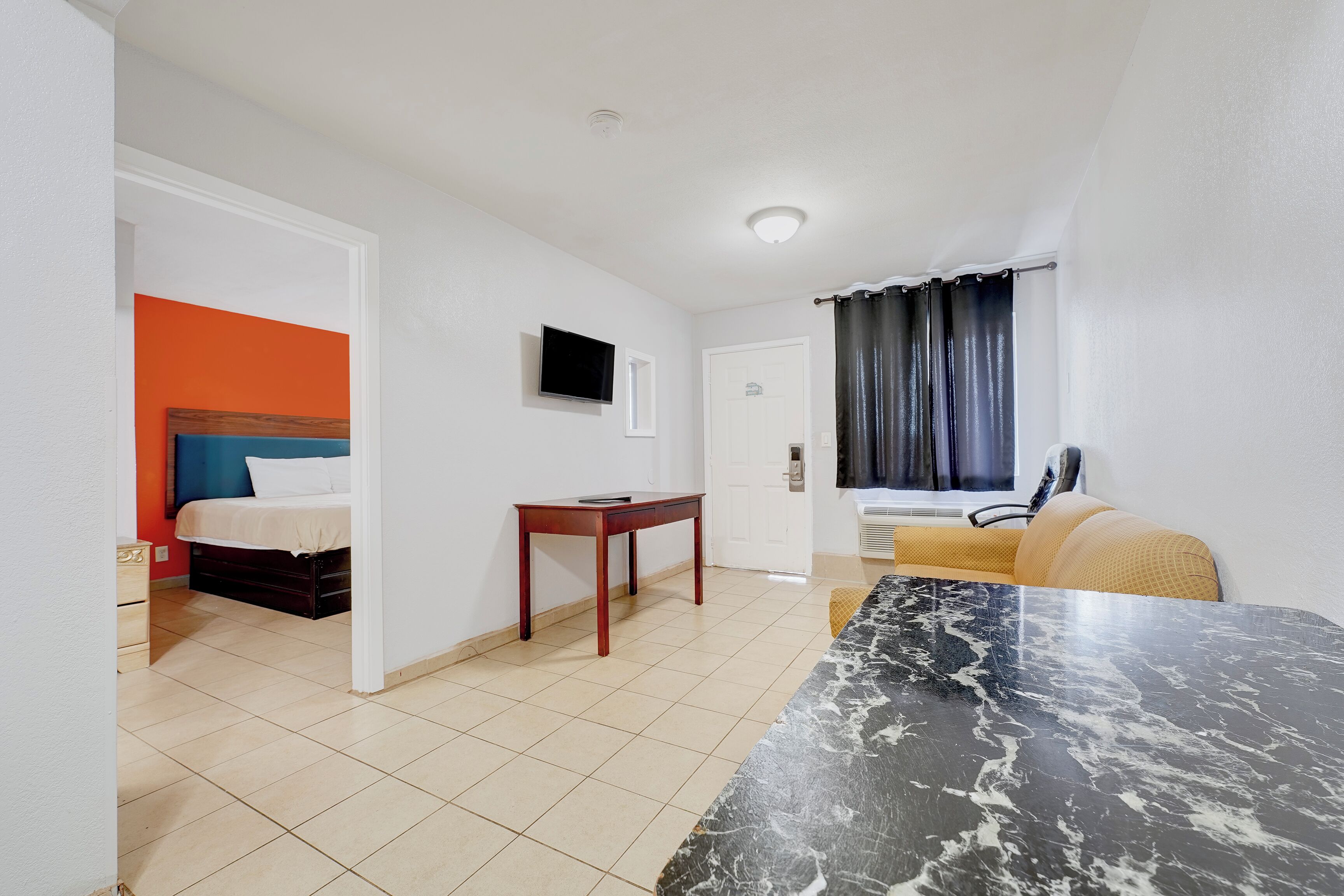 A hotel room with a bed, a small table, a TV, a window with curtains, and a tiled floor.