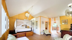 Quarto com uma cama, uma cadeira, uma mesinha e um ventilador.