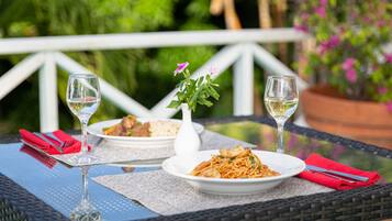 Una mesa de comedor con dos platos de comida y dos copas de vino, sobre un patio al aire libre con barandilla blanca y follaje al fondo.