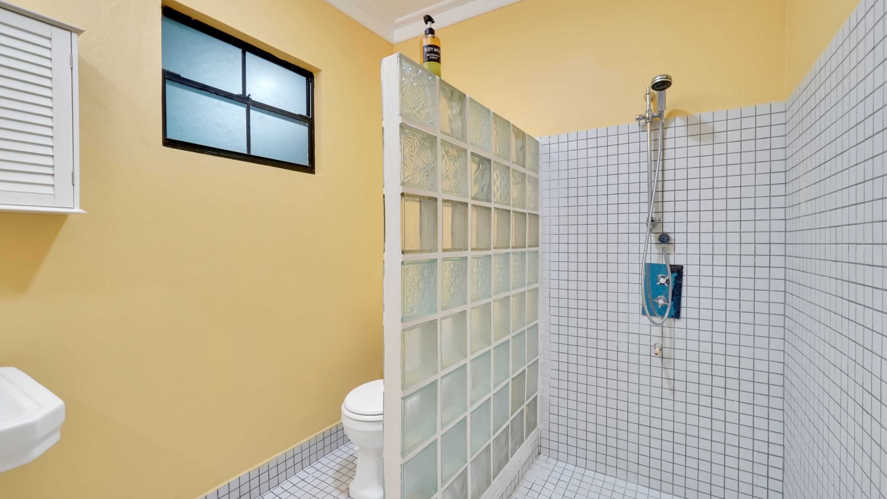 Un baño con inodoro, una ventana, ducha con cabezal de ducha manual y dispensador de jabón en la pared.