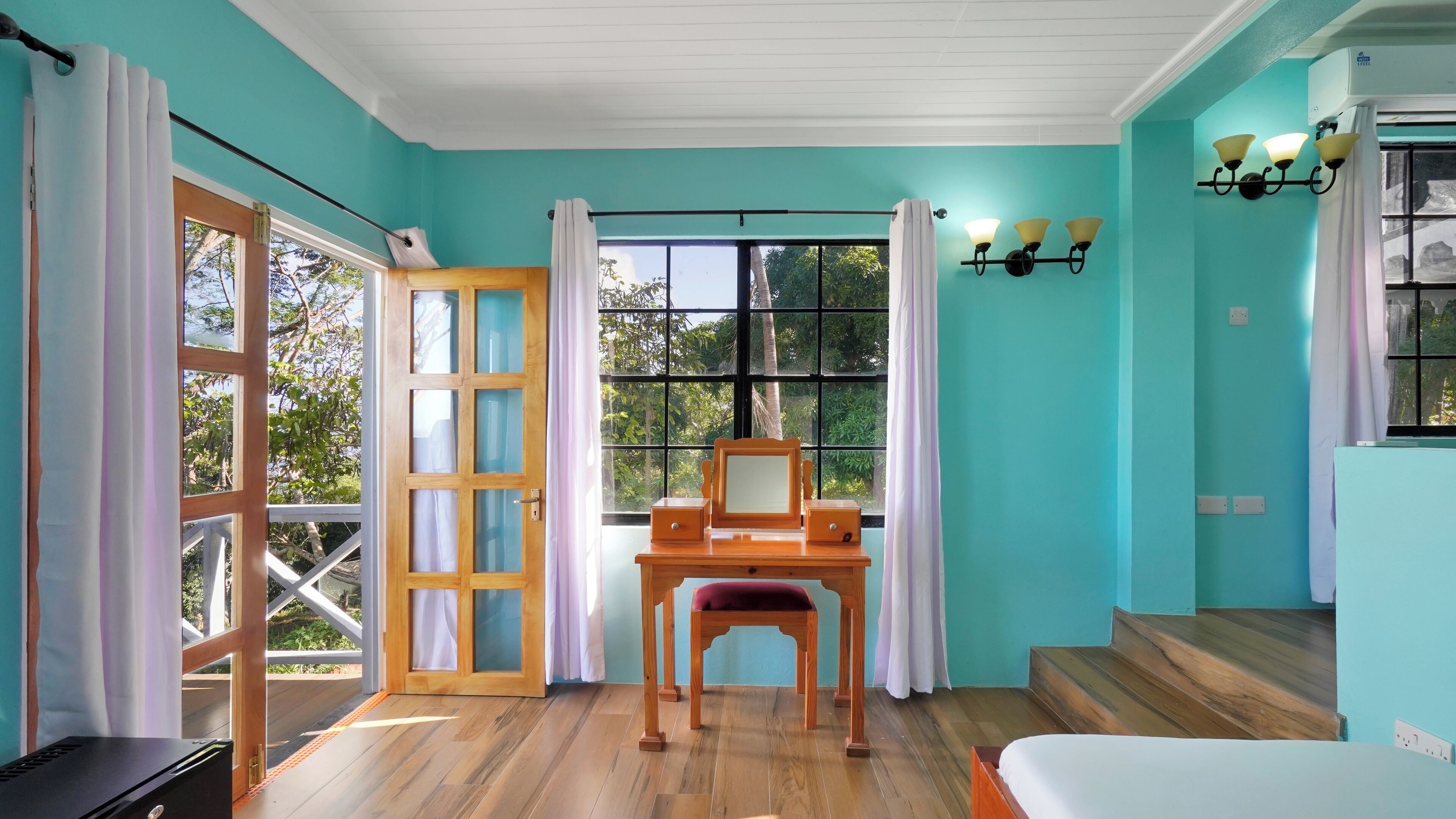 A room with teal walls, a wooden desk with a mirror, a bed, and a window with curtains.