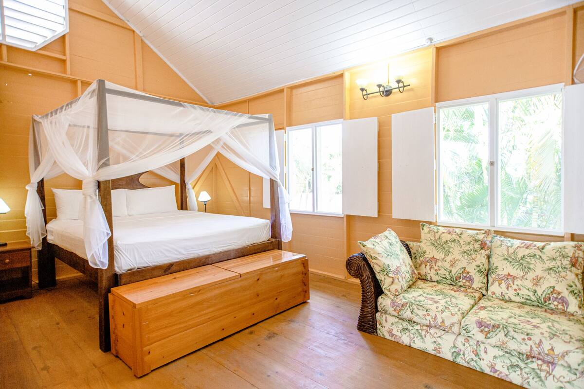 Quarto com cama de madeira com dossel, um banquinho, um sofá floral e uma janela com vista para o verde.