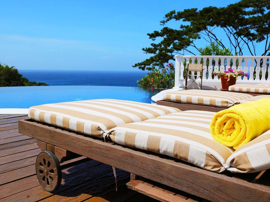 Une terrasse en bois avec une chaise longue et un transat au bord de la piscine, agrémentée de coussins à rayures et d’une serviette jaune.