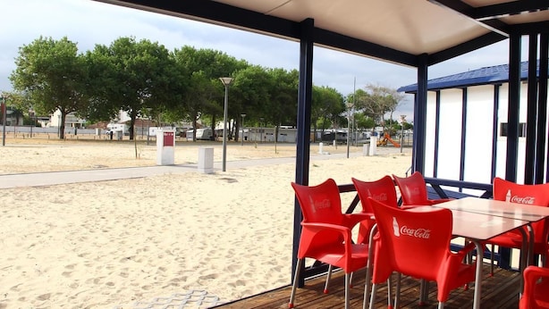 A beachside restaurant with red Coca-Cola branded chairs and tables, overlooking a sandy beach with trees and a clear sky. - Torreira Camping & Bungalows (Murtosa)