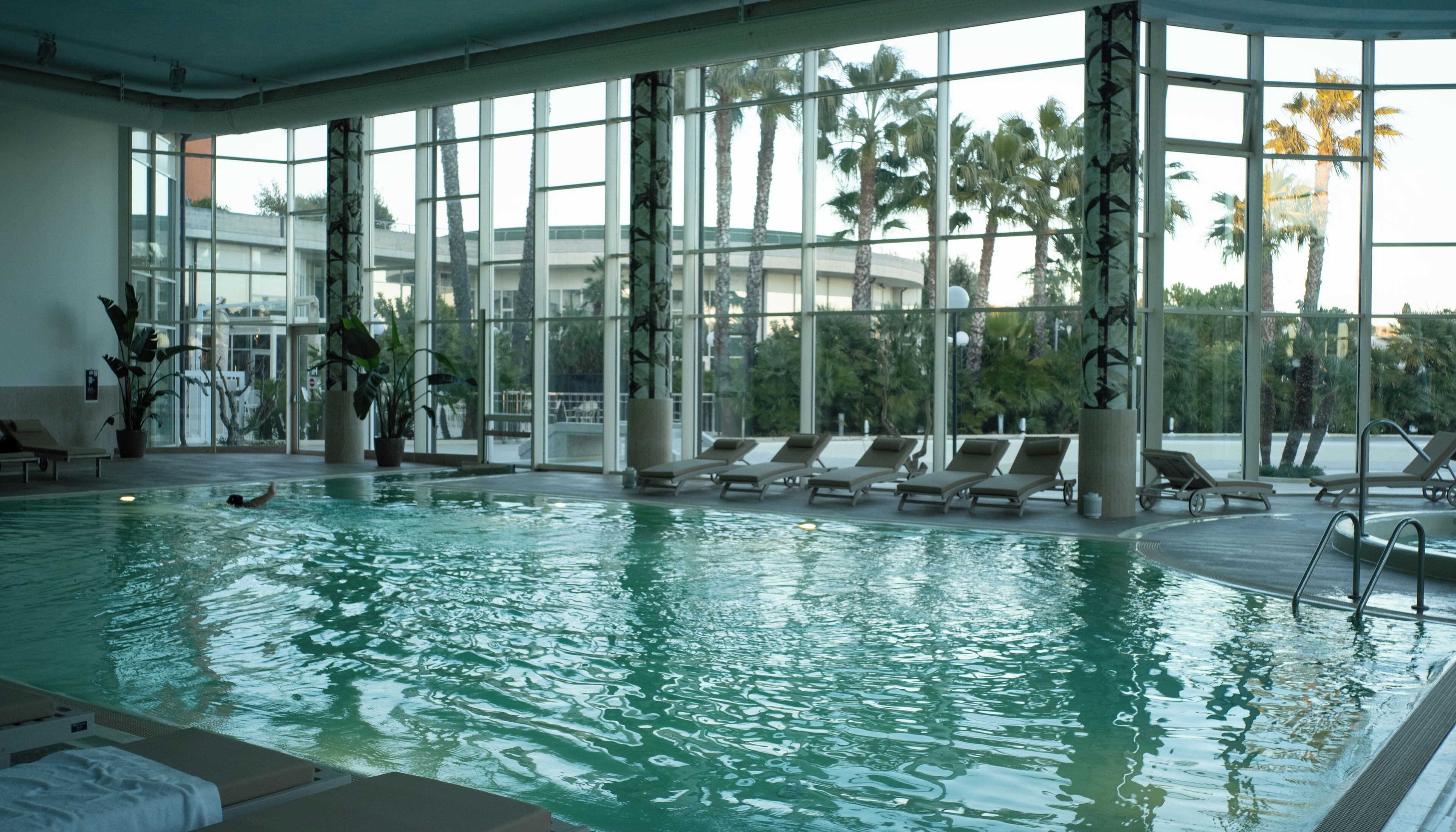 Una piscina interior con paredes de vidrio transparente, sillones de descanso y plantas en macetas.