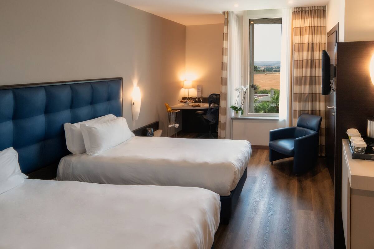 A hotel room with two beds, a desk with a lamp, a chair, a TV, and a window with a view.