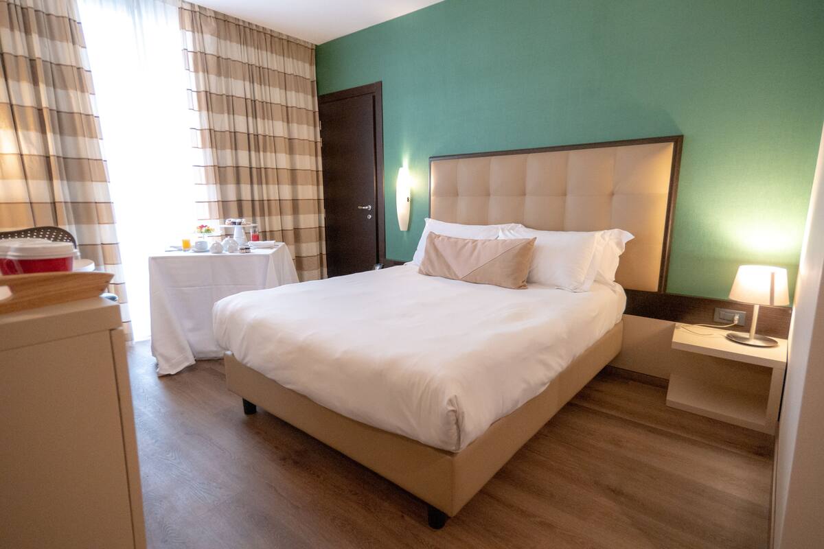 A hotel room with a large bed, a bedside table, a nightstand, a wardrobe, and a window with curtains.