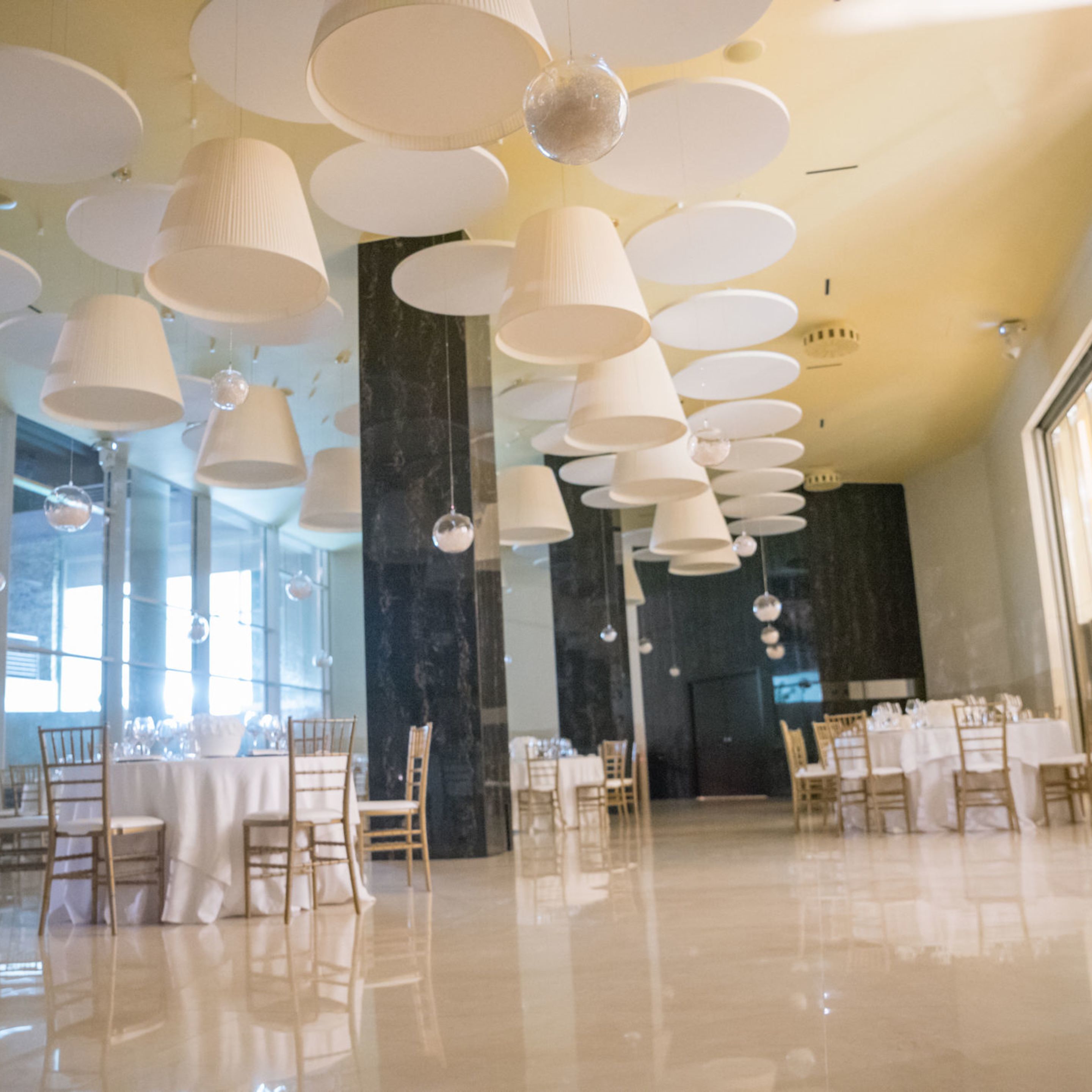 Un amplio salón de banquetes con varias mesas dispuestas para comer, grandes lámparas esféricas colgantes y un suelo pulido.