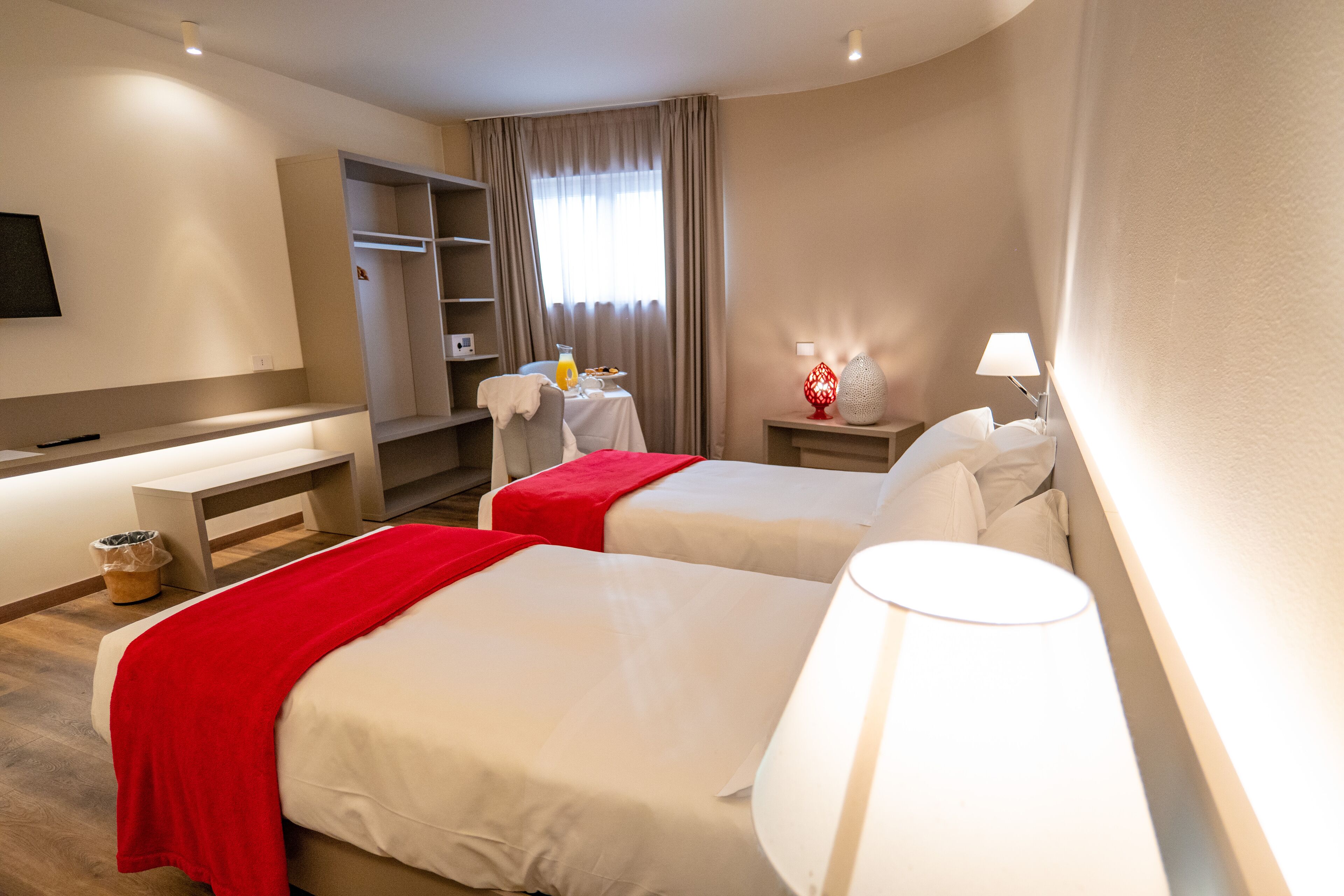 A hotel room with two beds, a desk, a chair, a TV, a lamp, a red decorative item, and a window with curtains.