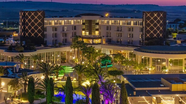 A hotel with a modern design, featuring a pool area, outdoor lighting, and a view of wind turbines in the background. - UNA HOTELS MH Matera (Borgo Venusio)