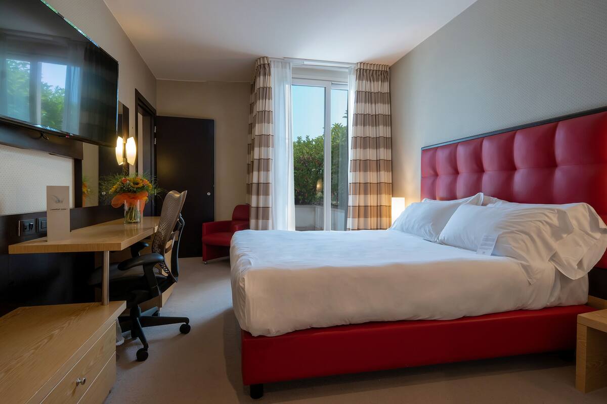 A hotel room with a large bed, a desk with a chair, a TV, and a window with curtains.